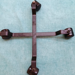 May include: A dark brown metal cross-shaped furniture base featuring four arms, each equipped with a small, dark brown caster wheel at the end. The structure is held together by a central metal fastener, and the arms have a flat, straight design.