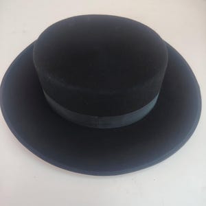 Op de afbeelding: Een effen zwarte boater-hoed met een platte kroon en een brede, ronde rand. De hoed is voorzien van een bijpassende zwarte grosgrain lintband die de basis van de kroon omsluit, wat zorgt voor een uniforme en monochrome uitstraling.