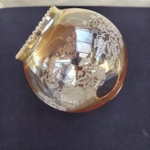 May include: A spherical amber-colored glass lamp shade featuring a white etched ivy leaf pattern. The opening at the top has a ruffled, scalloped edge. The glass has a reflective, iridescent finish that catches light across its curved surface.