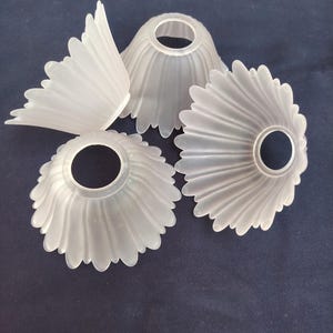 Pode incluir: Um conjunto de três cúpulas de lâmpada em vidro fosco, cada uma com uma borda recortada em forma de pétalas que lembra uma flor. O vidro translúcido tem um acabamento fosco e cada cúpula inclui uma abertura circular no topo para encaixe.