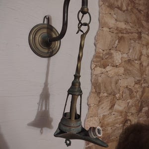 Peut inclure: Une applique murale en laiton composée d'un bras incurvé fixé à une plaque de base circulaire. Une tige métallique torsadée est suspendue au bras par un anneau, soutenant une structure en cage avec une douille d'ampoule sur le côté.