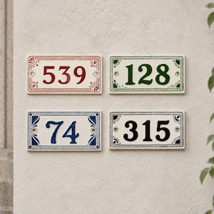 Könnte beinhalten: Vier rechteckige Hausnummernschilder aus Keramik mit abgerundeten Ecken und dekorativen Rändern. Das Schild oben links zeigt 539 in Rot, oben rechts 128 in Grün, unten links 74 in Blau und unten rechts 315 in Schwarz auf weißem Grund.