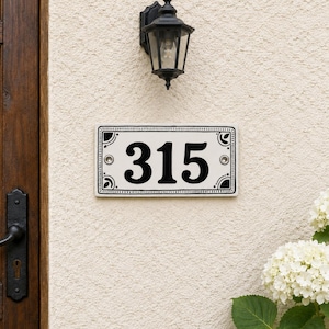 Könnte beinhalten: Ein rechteckiges Hausnummernschild aus Keramik mit den schwarzen Ziffern 315 auf weißem Grund. Das Schild hat einen dekorativen schwarzen Rand mit Eckverzierungen und ist mit zwei silbernen Schrauben an einer strukturierten beigen Wand befestigt.