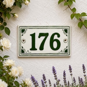 Peut inclure: Une plaque de numéro de maison rectangulaire en céramique affichant les chiffres 176 en vert foncé. La plaque présente un fond blanc avec une bordure vert foncé et des motifs décoratifs dans les coins. Deux trous de fixation sont visibles.
