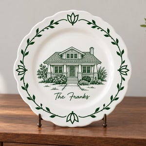 May include: A white ceramic plate with a scalloped edge featuring a dark green illustration of a house and the text The Franks. The border is decorated with a dark green vine and leaf pattern that encircles the central house illustration.