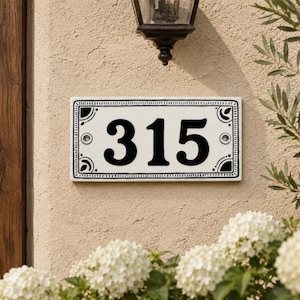 Könnte beinhalten: Ein rechteckiges Hausnummernschild mit den schwarzen Ziffern 315 auf weißem Grund. Das Schild hat einen dekorativen schwarzen Rand mit Eckverzierungen sowie zwei sichtbare Befestigungsschrauben auf der linken und rechten Seite.