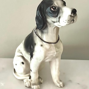 Op de afbeelding: Een keramieken beeldje van een zittende hond, overwegend wit met zwarte vlekken op zijn hoofd en rug. De hond heeft een bruine halsband en kijkt naar rechts. Een decoratief item.