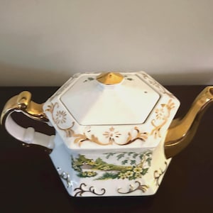 May include: A white teapot with a gold lid and handle. The teapot has a hexagonal shape and is decorated with gold trim and a floral design. The teapot is sitting on a dark surface.