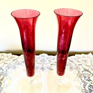 May include: Two tall, clear glass vases with a deep red, almost burgundy, color. The vases have a flared top and a clear glass stem with a round base.
