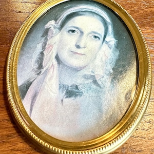 May include: Oval portrait miniature in a gold-colored frame. The portrait depicts a woman with a white head covering and a pink scarf. The frame has a decorative beaded edge. The portrait is set against a neutral background.