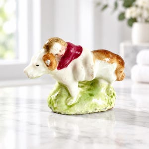 May include: A vintage ceramic figurine depicting a small child with light brown hair wearing a dark red garment, resting on the back of a white dog with brown patches. The entire piece is mounted on a textured, light green painted base.