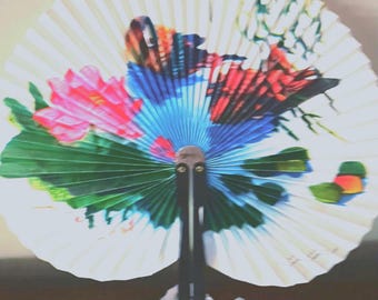 May include: A decorative hand fan with a floral and bird design. The fan features a white background with a colorful illustration of a pink flower, blue and red bird, and green leaves. The handle is black.