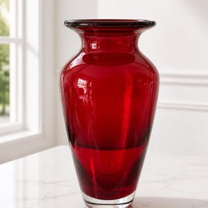 May include: A tall, translucent red glass vase with a wide, flared rim and a tapered neck that widens toward the center before narrowing at the base. The glass features a smooth, polished finish with a clear, thick base at the bottom.