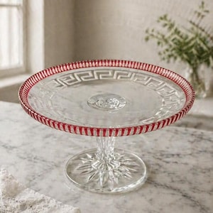 May include: A clear glass cake stand with a red and white patterned rim. The stand has a decorative base and a round top, perfect for displaying desserts. The stand is approximately 10 inches in diameter.