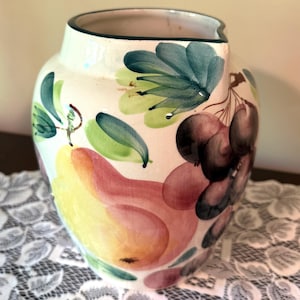 May include: A cream-colored ceramic pitcher featuring a hand-painted design of a large yellow and pink pear, clusters of dark purple grapes, and green leaves. The pitcher has a wide opening with a dark green rim and a rounded body shape.