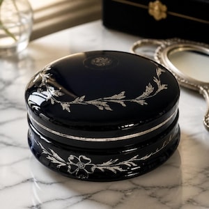 May include: A navy blue, round, two-tiered jewelry box with silver floral and leaf designs. The box has a glossy finish and silver trim. A small, silver-framed mirror and a black velvet box are in the background.