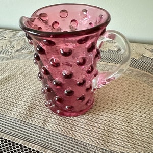 May include: A pink glass pitcher with a clear handle. The pitcher has a textured surface with small raised dots.