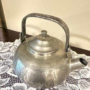 May include: A vintage, silver-colored metal teapot with a rounded body, a spout, and a large, arched handle. The lid has a small knob. The teapot sits on a white lace tablecloth.