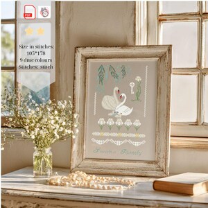 May include: A framed cross-stitch pattern featuring two white swans, a key, and floral motifs on a light background. The text reads "Swan Family". The design includes 9 DMC colors and measures 105 by 178 stitches. The frame is light-colored wood.