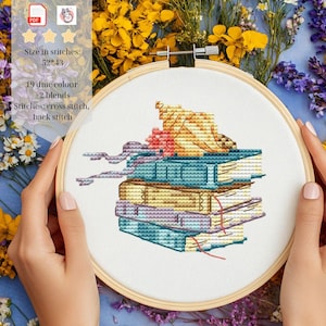 Pode incluir: Um padrão de ponto cruz num bastidor de madeira com uma pilha de quatro livros encimada por uma concha. O design inclui o texto: Size in stitches: 52*43, 19 dmc colour + 2 blends, Stitches: cross stitch, back stitch.