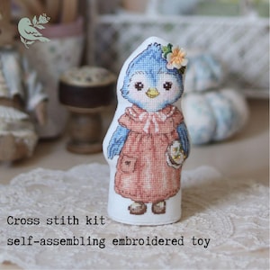 May include: A cross stitch kit for a self-assembling embroidered toy featuring a blue bird character wearing a peach-colored dress with a small flower accessory on its head. The text on the image reads: Cross stith kit self-assembling embroidered toy.