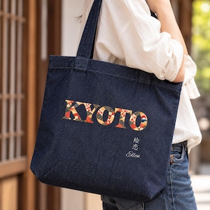 以下が含まれることがあります： 赤、白、金の小花模様で埋め尽くされた大きな大文字の「KYOTO」という文字が特徴のダークブルーのデニムトートバッグ。テキストの下には、「絵恋」という日本語の文字と「Ellen」という名前がプリントされています。