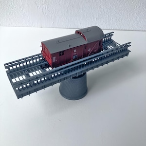 May include: A model train boxcar in dark red with a grey roof, featuring the letter W on its side, positioned on a grey plastic model railway bridge structure supported by a single central stone-textured pier base.
