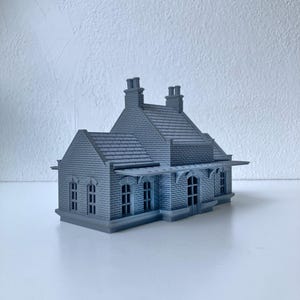 Può includere: Un modellino in miniatura monocromatico blu-grigio di una stazione ferroviaria caratterizzato da una dettagliata trama in mattoni, un tetto a tegole con due camini, finestre ad arco e una pensilina coperta lungo la facciata anteriore.