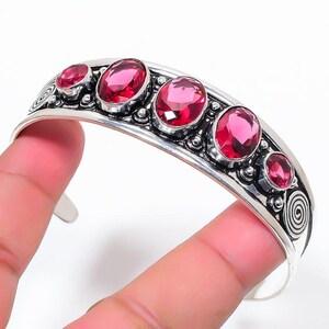 Pode incluir: Uma pulseira de prata tipo bracelete com cinco pedras preciosas cor-de-rosa de corte oval e facetadas dispostas em fila. A banda de metal é decorada com intrincados detalhes em prata e padrões em espiral nas extremidades, com textura.