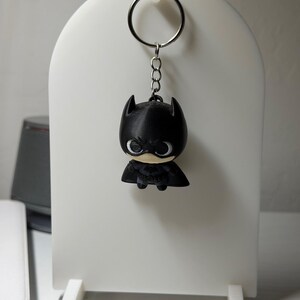 May include: A keychain featuring a small, stylized black Batman figure with a large head, pointed ears, and a cape. The figure is attached to a silver-colored metal split ring by a short chain, showcasing a matte finish and detailed facial features.
