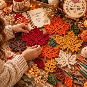 May include: A collection of crocheted autumn leaves in shades of red, orange, yellow, brown, and cream, arranged alongside a notebook with the text Handmade with Love, a wooden embroidery hoop with the text Corner Store PK, and metal scissors.