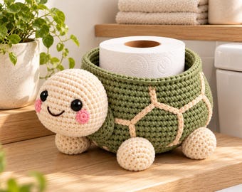 May include: A crochet turtle-shaped toilet paper holder featuring a green shell with cream-colored stitching, a cream-colored head with black eyes and pink cheeks, and four small cream-colored feet. The shell holds a roll of white toilet paper.