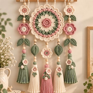 May include: A wall hanging featuring a large central crochet mandala in cream, pink, and sage green. It is suspended from a wooden dowel with hanging strands of crochet flowers, green leaves, wooden beads, and long tassels in pink and cream.