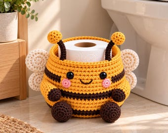 May include: A crocheted bee-shaped toilet paper holder featuring a yellow and brown striped body, two antennae with round tips, small arms, and white wings. The round container has a smiling face with pink cheeks and black eyes.