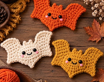 May include: Three crocheted bat ornaments in orange, cream, and mustard yellow. Each bat features black safety eyes, a stitched smile, and pink cheeks. The bats are arranged on a wooden surface alongside yarn balls and a crochet hook.