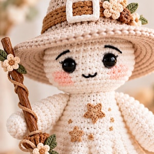 May include: A crocheted doll wearing a beige pointed hat with a brown band, a white buckle, and small fabric flowers. The doll has black eyes, pink cheeks, and a white dress decorated with small brown stars. It holds a brown broom with flowers.