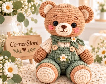 May include: A crocheted teddy bear with light brown fur, black eyes, and pink cheeks. The bear wears cream-colored sleeves and sage green overalls featuring two wooden buttons and a small white flower appliqué on the front pocket.