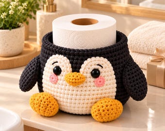 May include: A black and white crocheted penguin-shaped toilet paper holder with a cream-colored belly, orange feet and beak, and pink cheeks. The penguin features two black plastic eyes and holds a standard roll of white toilet paper.