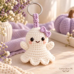 May include: A white crocheted ghost keychain featuring a small purple bow, black bead eyes, a stitched smile, and pink blush accents. The ghost is attached to a silver metal keyring by a purple crocheted loop. The text Corner Store PK is visible.