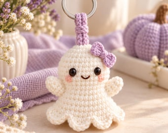 May include: A white crocheted ghost keychain featuring a small purple bow, black bead eyes, a stitched smile, and pink blush accents. The ghost is attached to a silver metal keyring by a purple crocheted loop. The text Corner Store PK is visible.
