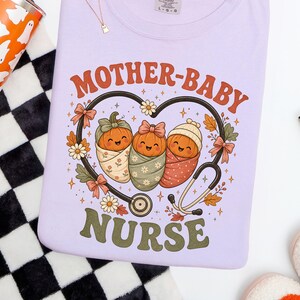 May include: A light purple t-shirt featuring a graphic of three swaddled pumpkins inside a heart-shaped stethoscope. The text reads MOTHER-BABY NURSE. The design includes small flowers, leaves, and bows in orange, green, and white colours.