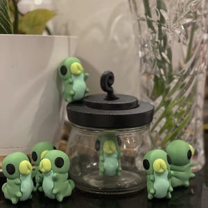 Peut inclure: Un bocal en verre transparent avec un couvercle noir et une poignée en spirale, entouré de plusieurs petites figurines de chenilles vert clair. Chaque figurine présente un ventre vert pâle, une tête arrondie, de grands yeux noirs et une petite bouche jaune clair.