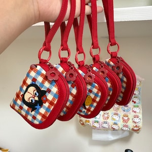 Puede incluir: Cinco estuches rectangulares pequeños con cremallera y estampado de cuadros rojos, blancos y azules. Cada estuche tiene una correa de muñeca roja sujeta por una anilla roja y un tirador de cremallera en forma de flor roja.