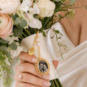 Può includere: Un ciondolo in metallo dorato per bouquet da sposa con una cornice ovale contenente una fotografia in bianco e nero di una coppia, una piccola targhetta rotonda incisa con il testo "I'm always with you" e una catenina dorata con due perle.