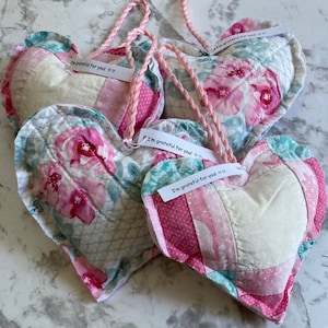 以下が含まれることがあります： 「あなたに感謝しています」というメッセージが書かれた、ピンクと白のパッチワークのハート型のクッションが4つあります。