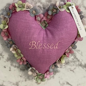 Könnte beinhalten: Ein rosa herzförmiges Kissen mit dem weißen Stickwort "Blessed" und einem floralen Besatz. Ein kleines Schild, das an das Kissen befestigt ist, lautet "Ich bin dankbar für dich".