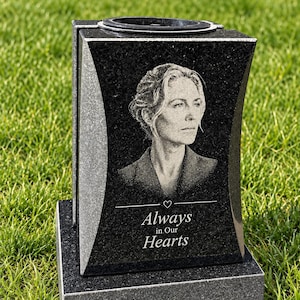 Pode incluir: Um vaso memorial de granito preto com uma superfície frontal polida apresentando um retrato gravado a laser de uma pessoa e o texto "Always in Our Hearts" com um pequeno ícone de coração. A pedra retangular tem lados curvos e uma base.