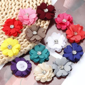 Puede incluir: Una colección de adornos de flores de tela multicolor, cada una con pétalos en capas y un acento central de diamantes de imitación transparentes. El conjunto incluye flores rojas, rosas, amarillas, marrones, blancas, azul verdoso, azules, moradas, beige y grises.