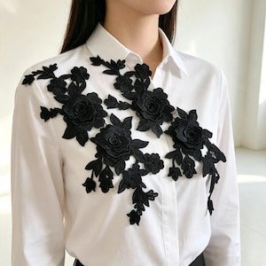 Può includere: Una camicia bianca con colletto abbottonato caratterizzata da un intricato ricamo floreale nero sul petto. Il design è composto da dettagliati motivi di rose nere con foglie e steli, applicati come un'applicazione che va dalla spalla al centro.
