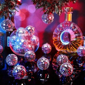 May include: A collection of small, spherical disco ball ornaments covered in reflective silver mirror tiles. Several ornaments are contained within a clear glass bowl, while others are scattered around a large, clear glass decanter.